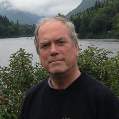 Man standing outdoors by a lake with mountains and trees in the background.